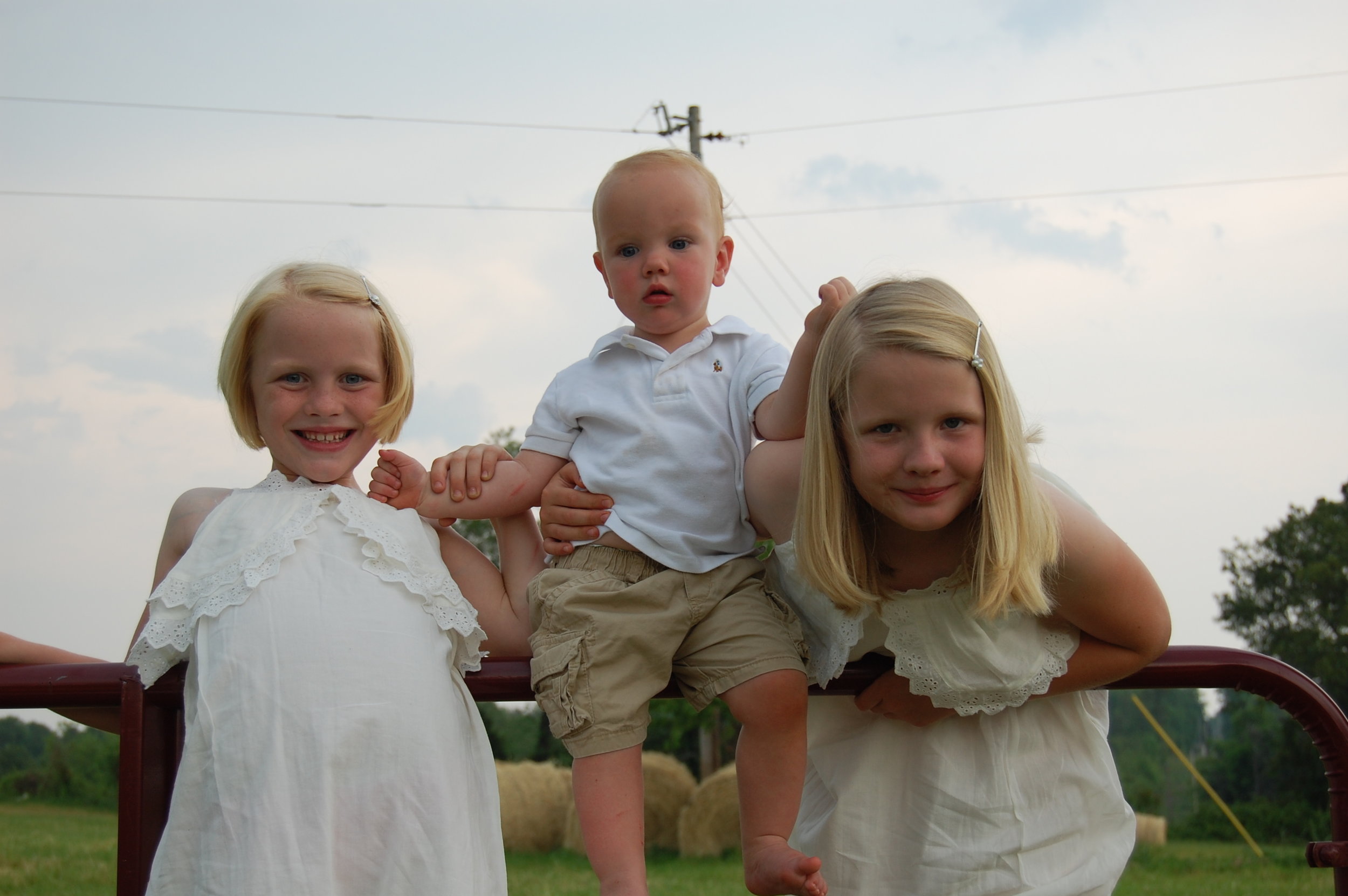 DSC_0624 Three young children, two girls and a boy, pose together outdoors. The girls wear white dresses, while the boy is in a white polo shirt and beige shorts. They are smiling and holding onto each other, with a grassy field and hay bales in the background under a cloudy sky.