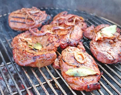 Ella's+Pastured+Pork Grilled marinated steaks cooking on a barbecue grill, featuring grill marks and garnished with onions and bay leaves, surrounded by smoke.