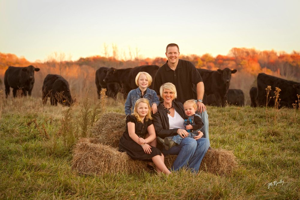family Family portrait on a farm with two children and a toddler sitting on hay bales, surrounded by black cows in a picturesque autumn setting. The background features colorful fall foliage, creating a warm and inviting rural atmosphere.